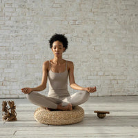 Femme en tenue de yoga beige, assise en position de méditation sur un coussin en osier, dans une pièce avec un mur en briques blanches et un sol en bois clair, avec des statues et un bol tibétain à côté.