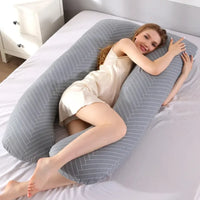 Femme allongée sur un lit, étreignant un grand coussin de grossesse gris avec des motifs à rayures blanches, dans une chambre lumineuse avec une table de chevet blanche.