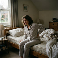 Une femme d'âge moyen en pyjama gris assise sur le bord de son lit, grimaçant de douleur et tenant le bas de son dos avec une main.