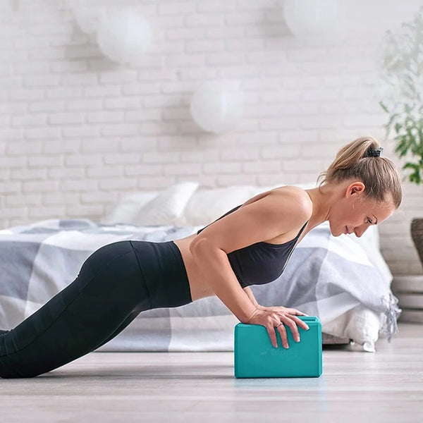 Coussin de yoga en mousse vert une femme s'aide de se cube pour faire des exercices de sport derrière elle un lit blanc et un mur de brique blanc