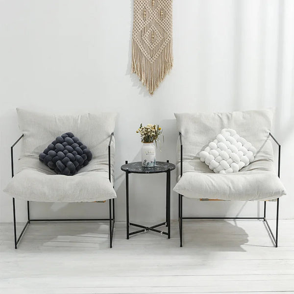 Coussin canapé en tricot noir et blanc dans un intérieur blanc, sur un fauteuil à gauche posé le coussin canapé en tricot noir et à côté sur un autre fauteuil posé le coussin canapé en tricot blanc avec entre les deux une table basse et un vase contenant des fleurs posé dessus
